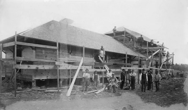 Carpenters on scaffolding pose in front of a large hip-roofed wood building under construction