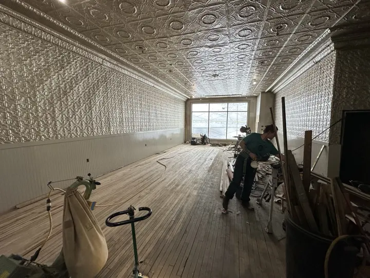 A worker refinishing the original maple floor beneath the restored tin walls and ceiling