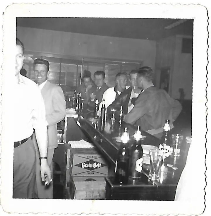 Men in suits and jackets lined up along the bar with bottles of beer and a case of Grain Belt on the counter