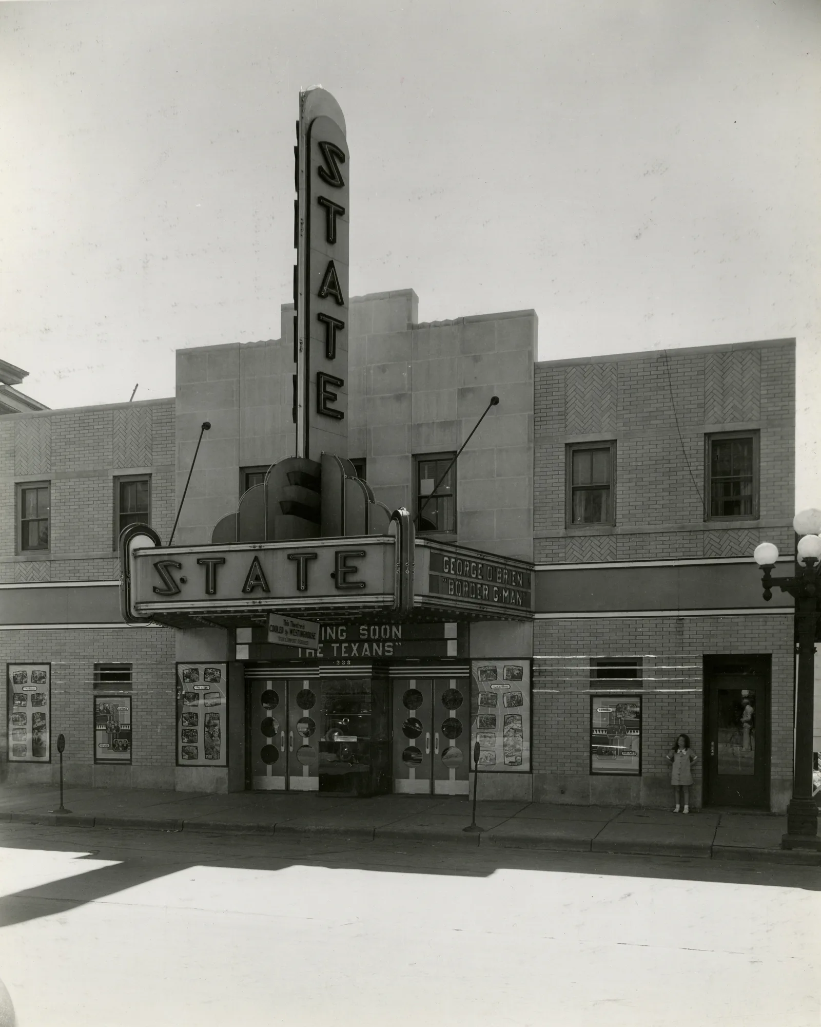 The State Theater around 1938 with its tall STATE sign, neon marquee advertising George O'Brien in Border G-Man and Coming Soon The Texans, and a young girl standing by the wall