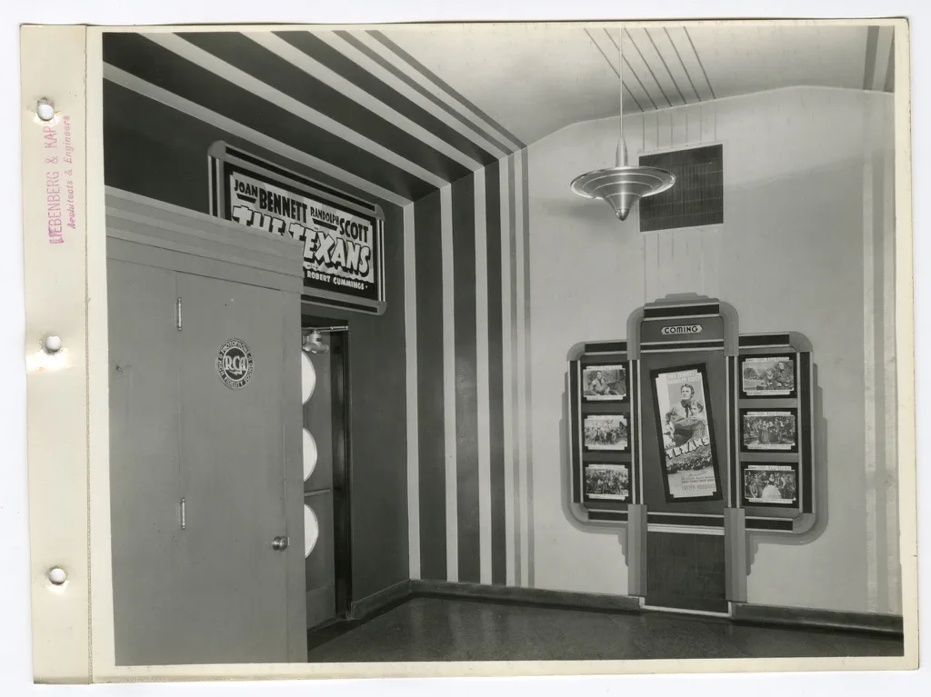 Theater lobby with bold striped walls, a saucer-shaped pendant light, a coming attractions frame and a poster for The Texans
