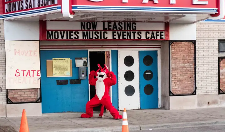 The mascot flexing beneath a marquee reading Now Leasing, Movies Music Events Cafe