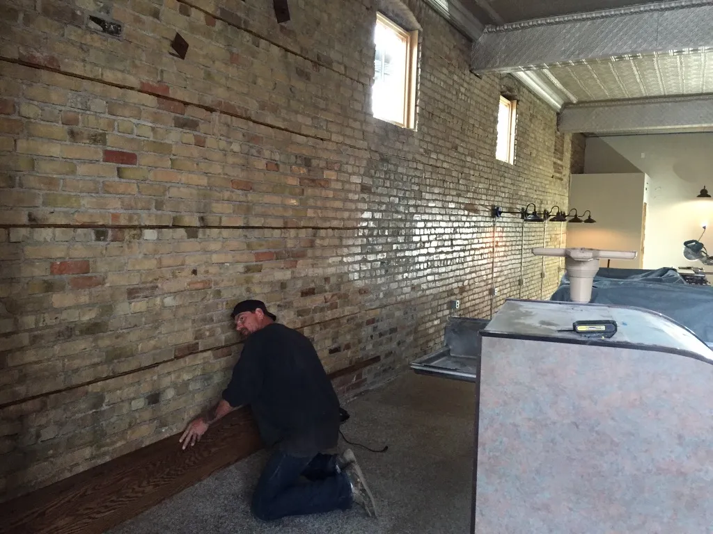A carpenter fitting wood trim along an exposed brick wall
