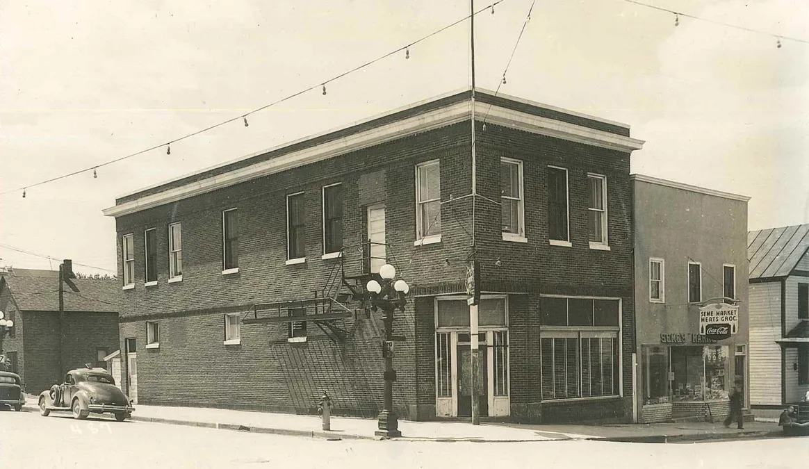 The two-story brick building on the corner in an early photo, with a globe street light, a fire escape, a vintage car and the Seme Market next door